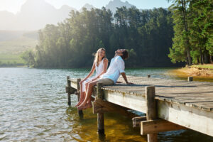 Young couple, lake and jetty with laughing, happiness and bonding with love in nature for holiday. Man, woman and comic joke on bridge by water to relax with conversation, care and vacation in summer.